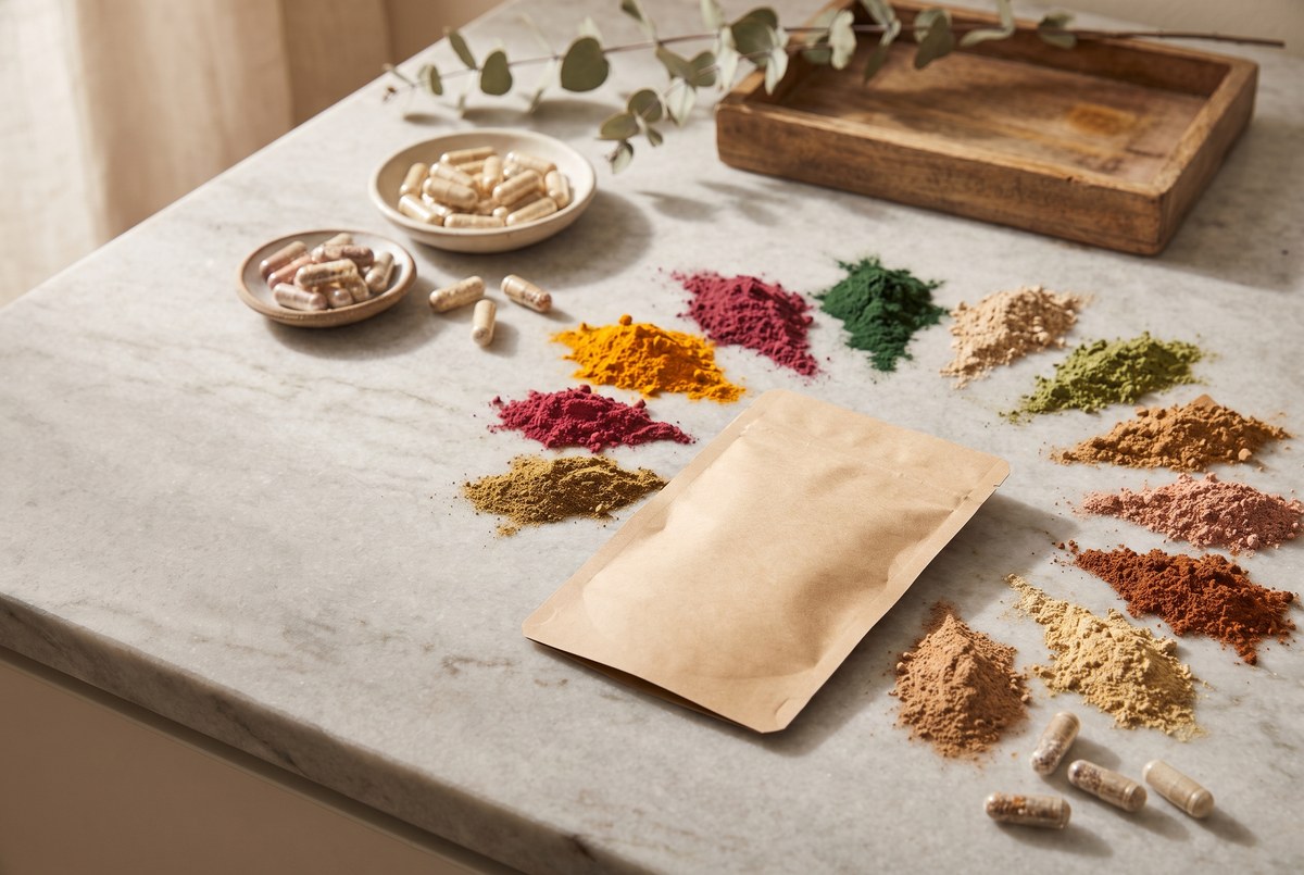 A single kraft-paper sachet beside twelve small piles of colorful powders and capsules fanned out on a marble counter, warm light, photographed for Magellan Longevity's review of twelve ingredients, one sachet, and the trial nobody has run.