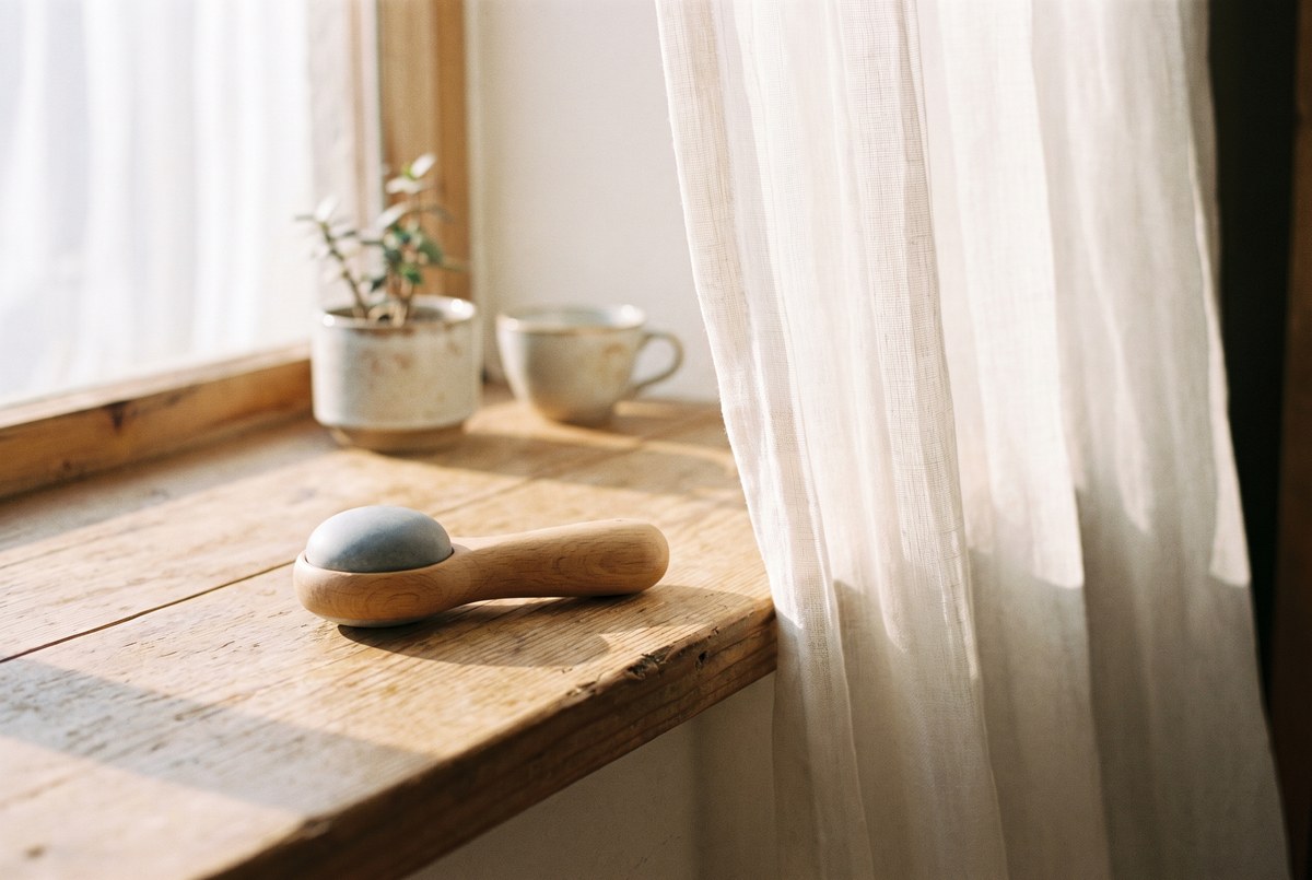 A small handheld breathing-resistance trainer resting on a sunlit wooden windowsill beside a softly billowing sheer curtain, calm morning light, photographed for Magellan Longevity's review of thirty breaths against resistance: the strangest blood-pressure trial in the longevity aisle.