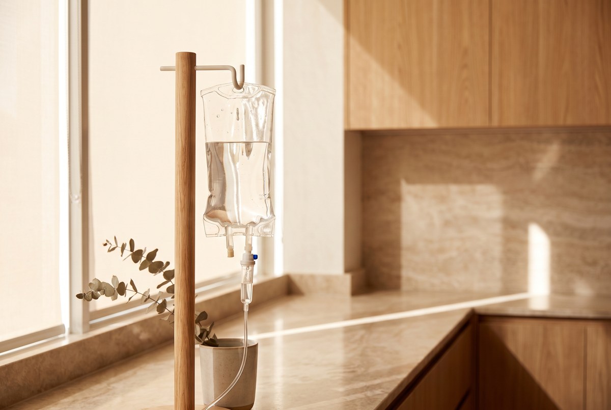 A glass IV drip bag with clear fluid hanging softly lit in a clean modern clinic room with wood accents, warm light, photographed for Magellan Longevity's review of iv nad+ clinics are selling ahead of the clinical evidence.