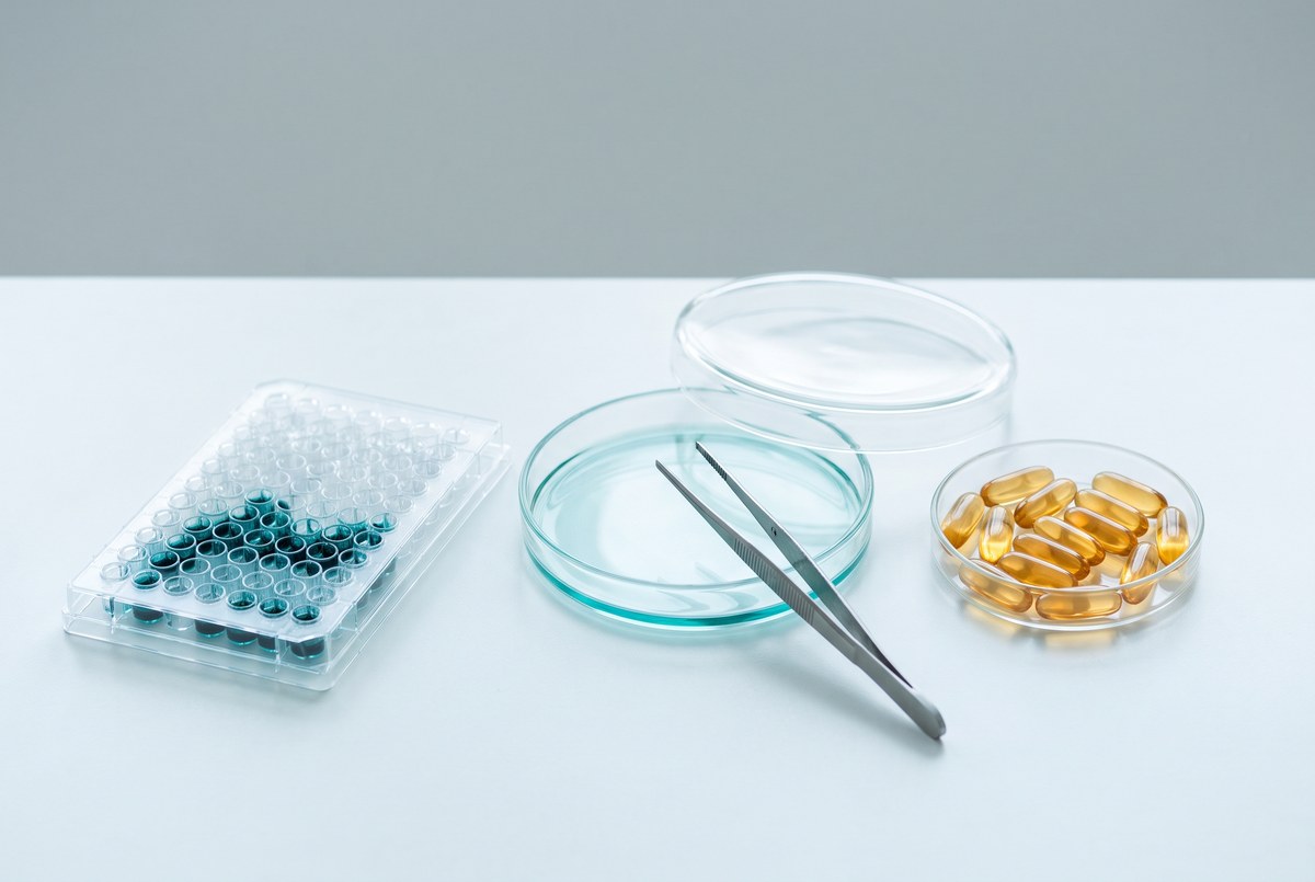 A bright laboratory bench holding a 96-well microplate with several wells stained blue-green, two glass petri dishes with fine steel forceps, and a small glass dish of amber capsules, photographed for Magellan Longevity's evidence review of senolytic supplements.